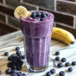 Close-up of a creamy Blueberry Banana Smoothie in a glass with blueberries and banana slices around it.