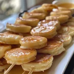 A close-up side view of mini pancakes stacked on skewers, drizzled with syrup.