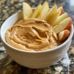 Close-up side view of a creamy peanut butter yogurt dip in a bowl, with sliced apples surrounding it.