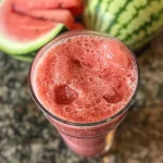 A close-up view of a vibrant watermelon coconut smoothie in a glass, garnished with a slice of watermelon.