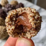 ['Close up of golden brown quinoa bites with a glossy chocolate coating and a sprinkle of sea salt.', 'Side view of crispy quinoa snacks with a rich dark chocolate exterior and visible quinoa texture.', 'Juicy quinoa bites coated in dark chocolate, highlighting their crunchy and chewy appearance.', 'Textured quinoa balls glistening with chocolate and topped with sea salt, displayed on a wooden surface.']