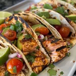 ['Close-up view of grilled salmon tacos topped with avocado salsa, served in a tortilla.', 'Juicy grilled salmon tacos with fresh avocado salsa, garnished with lime wedges and herbs.', 'A side view of vibrant grilled salmon tacos filled with diced avocados and crunchy cabbage.', 'Colorful presentation of grilled salmon tacos with avocado salsa and cherry tomato garnish.']