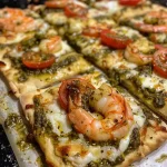 ['Close-up view of a Pesto Shrimp Flatbread topped with cherry tomatoes and cheese.', 'Side view of a Pesto Shrimp Flatbread with vibrant ingredients and melted cheese.', 'Juicy Pesto Shrimp Flatbread showcasing shrimp, pesto, and colorful toppings.', 'Deliciously garnished Pesto Shrimp Flatbread with shrimp and fresh basil.']