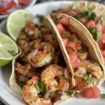 ['A close-up side view of juicy shrimp tacos topped with colorful watermelon pico de gallo.', 'Shrimp tacos filled with fresh ingredients, served with vibrant watermelon salsa.', 'Side view showing delicious shrimp tacos garnished with diced watermelon and cilantro.', 'An appetizing display of shrimp tacos accompanied by bright watermelon pico de gallo.']