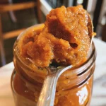 ['A close-up view of creamy pumpkin butter in a glass jar.', 'A richly colored pumpkin butter spread with a smooth texture on a wooden board.', 'Pumpkin butter with a glossy finish, displayed in a bowl with a spoon.', 'A side angle of thick pumpkin butter, highlighting its vibrant orange hue.']