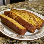 ['Close-up view of freshly baked yellow squash bread with a golden crust.', 'Slice of yellow squash bread showing moist interior and garnished with herbs.', 'Side view of a loaf of yellow squash bread on a wooden cutting board.', 'A piece of yellow squash bread displayed on a plate, highlighting its texture.']