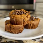 ['Close-up side view of moist pumpkin protein muffins topped with mini chocolate chips.', 'Delicious pumpkin protein muffins on a wooden table, showcasing a soft and fluffy texture.', 'A freshly baked pumpkin protein muffin with a golden crust and chocolate chips, captured from the side.', 'Side view of pumpkin protein muffins, highlighting their rich color and inviting appearance.']
